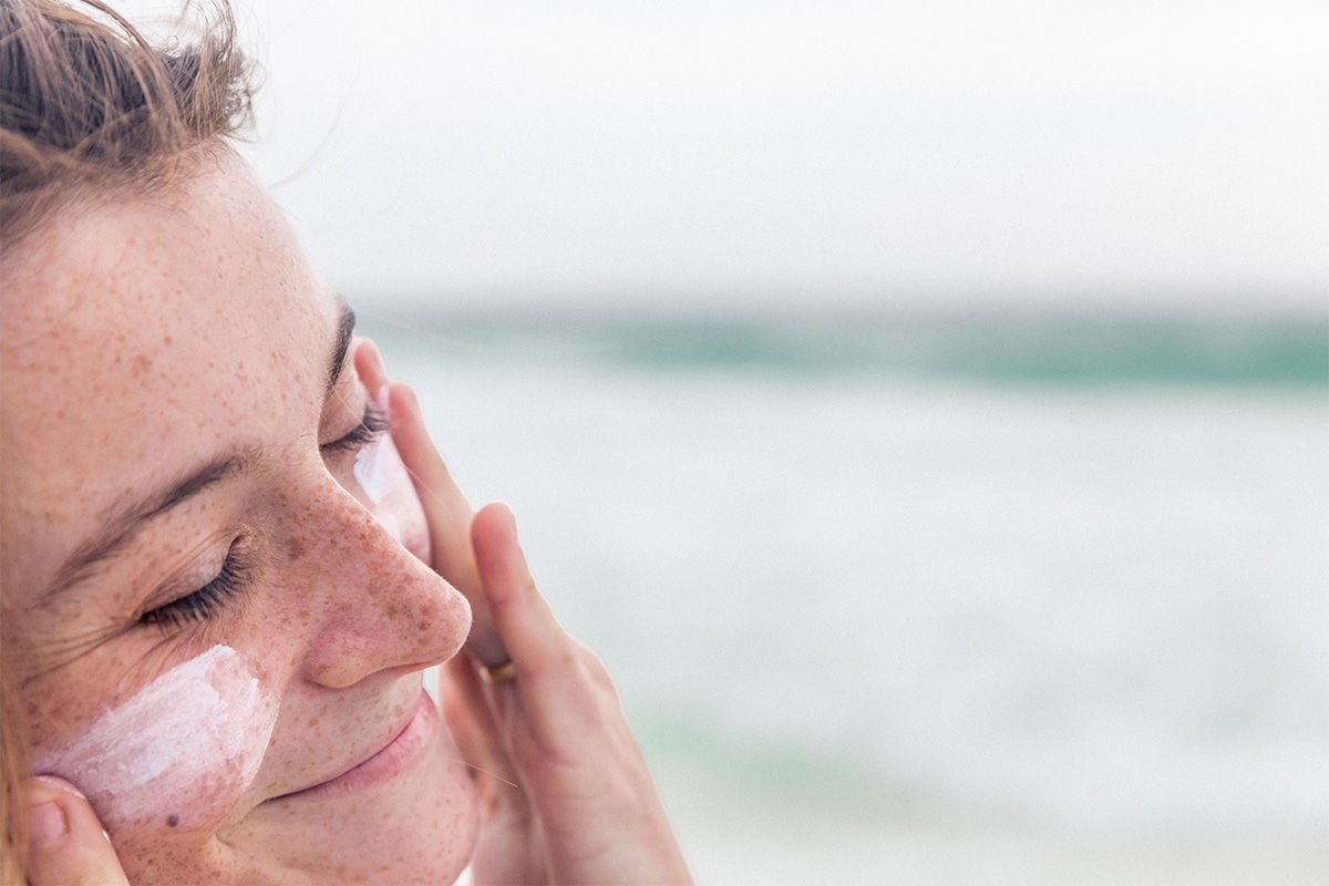 A woman applies sunscreen to her face at the beach. 